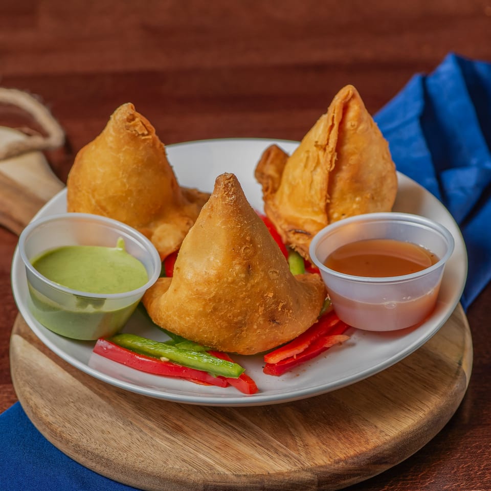 Three golden-fried Milagu Indian Cuisine samosas on a white plate with green and red peppers, and two dipping sauces.
