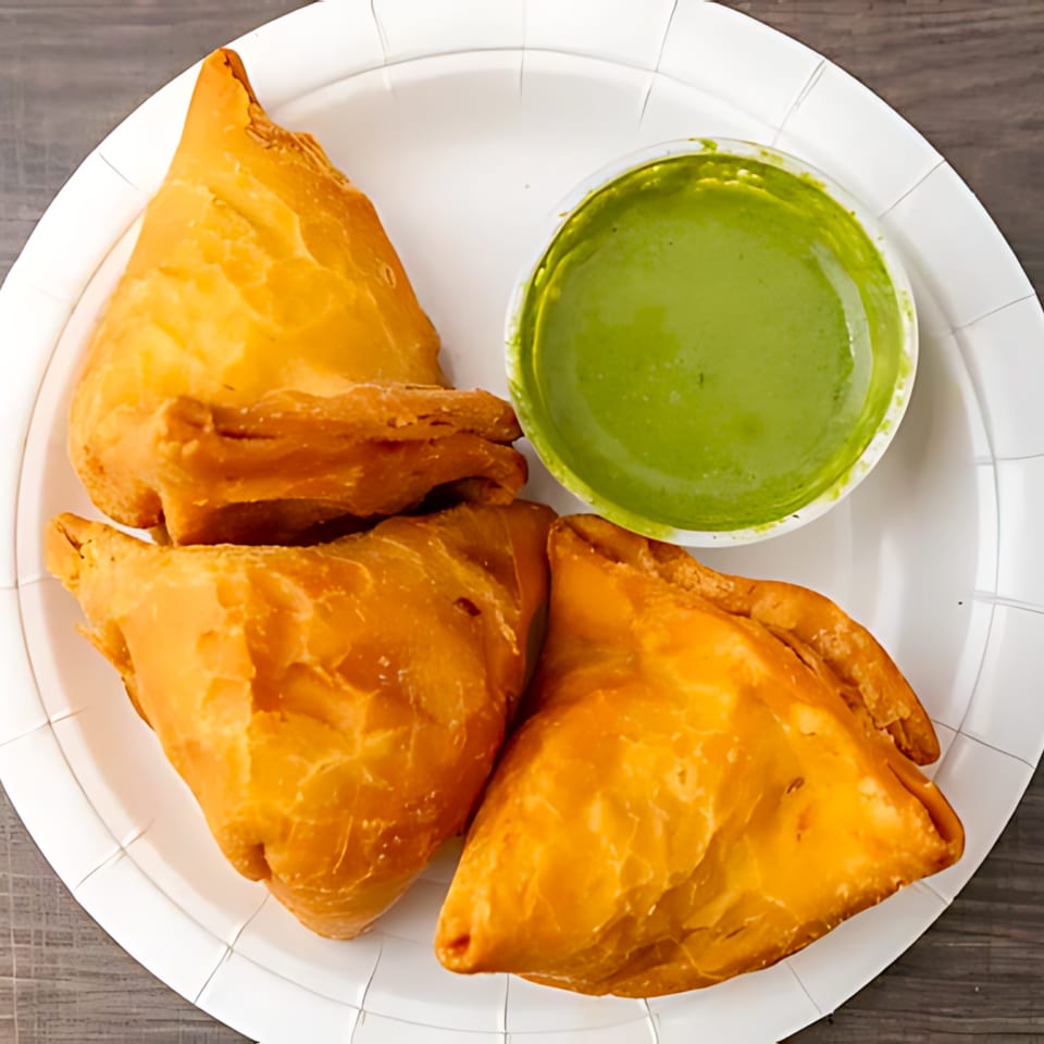 Three golden-brown samosas and a small cup of vibrant green chutney on a white paper plate.